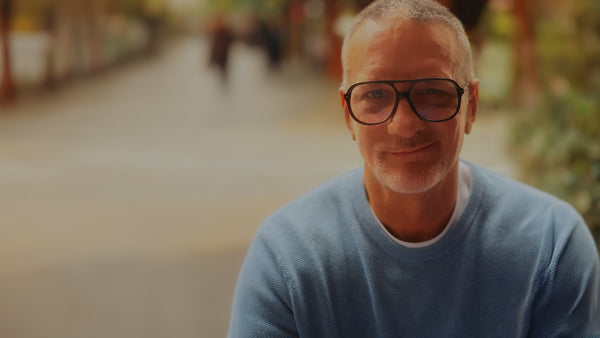 A close-up portrait of a man with short gray hair and glasses, wearing a light blue knit sweater over a white undershirt, set against a softly blurred outdoor background.